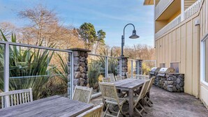 An outdoor patio with a stone wall, wooden tables and chairs, a grill, and a metal railing.