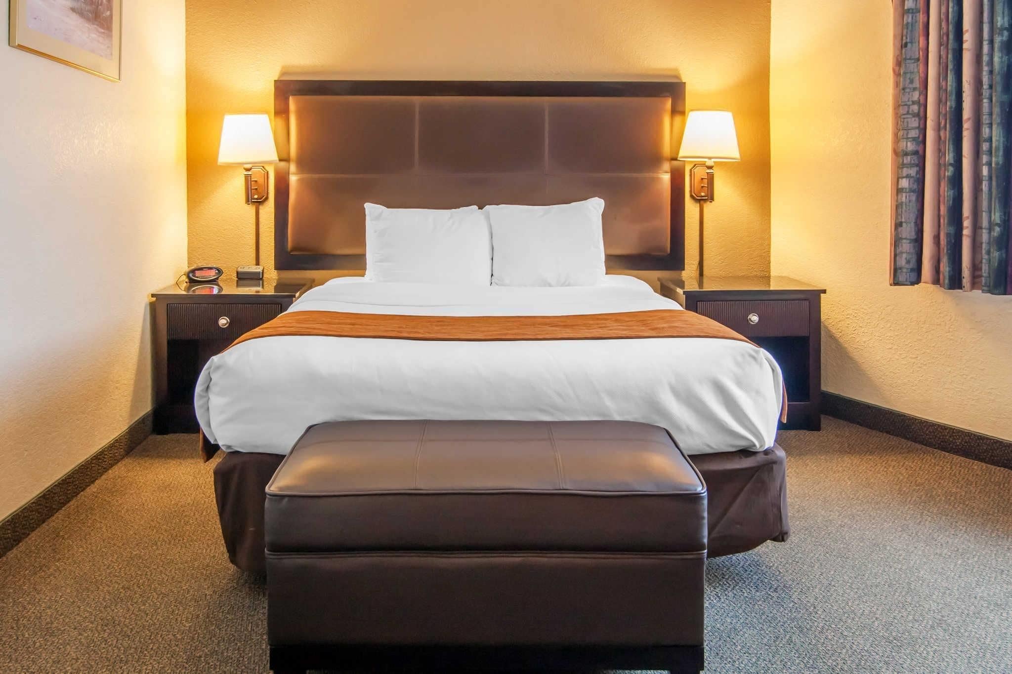 A hotel room with a large bed, two bedside lamps, a nightstand, and a brown leather ottoman.