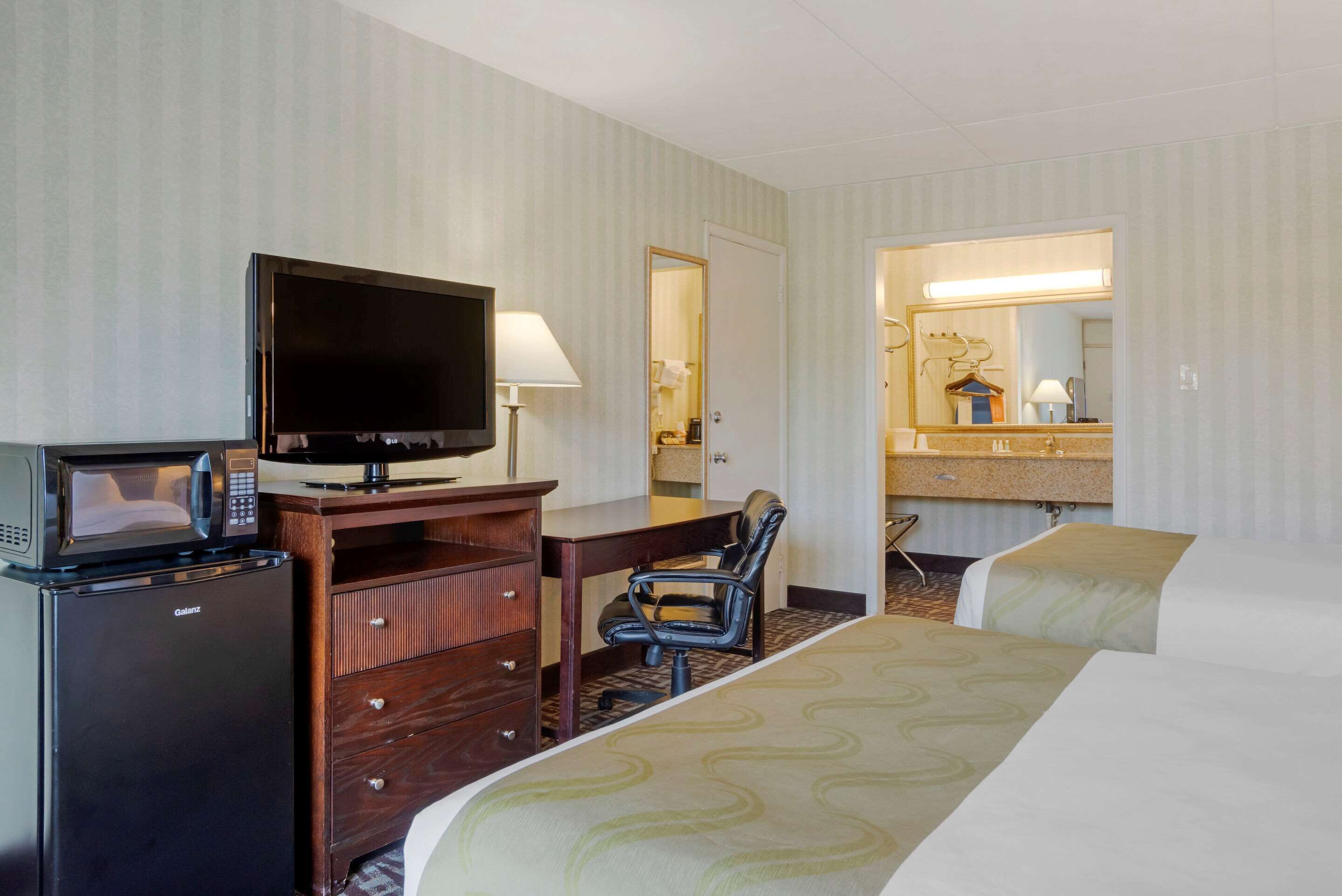Habitación de hotel con cama, escritorio con televisor, microondas, silla y baño visible a través de una puerta abierta.