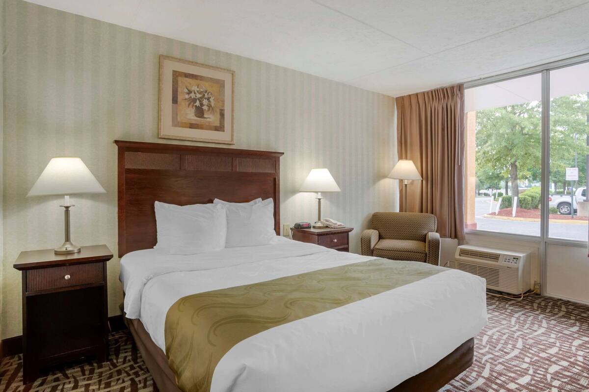 Ein Hotelzimmer mit einem Bett, zwei Nachttischen mit Lampen, einem Sessel, einem Fenster mit Vorhängen und einer Wandklimaanlage.