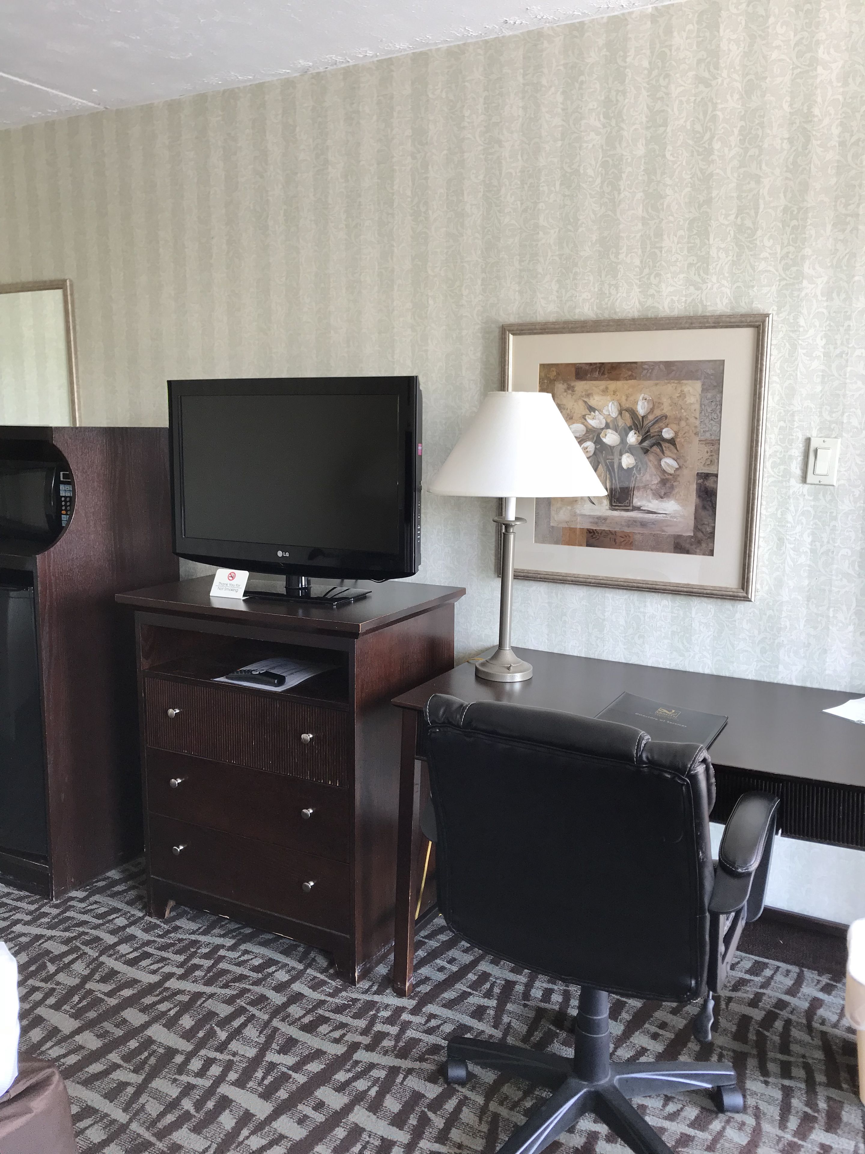 Een hotelkamer met een bureau, een televisie, een lamp, een lijst met een afbeelding en een kamerbreed tapijt.