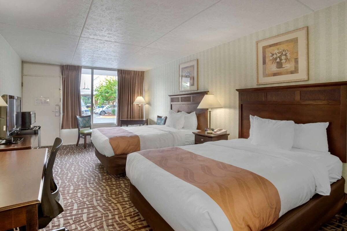 Ein Hotelzimmer mit zwei Betten, einem Schreibtisch, einem Stuhl, einem Fernseher und einem Fenster mit Vorhängen.