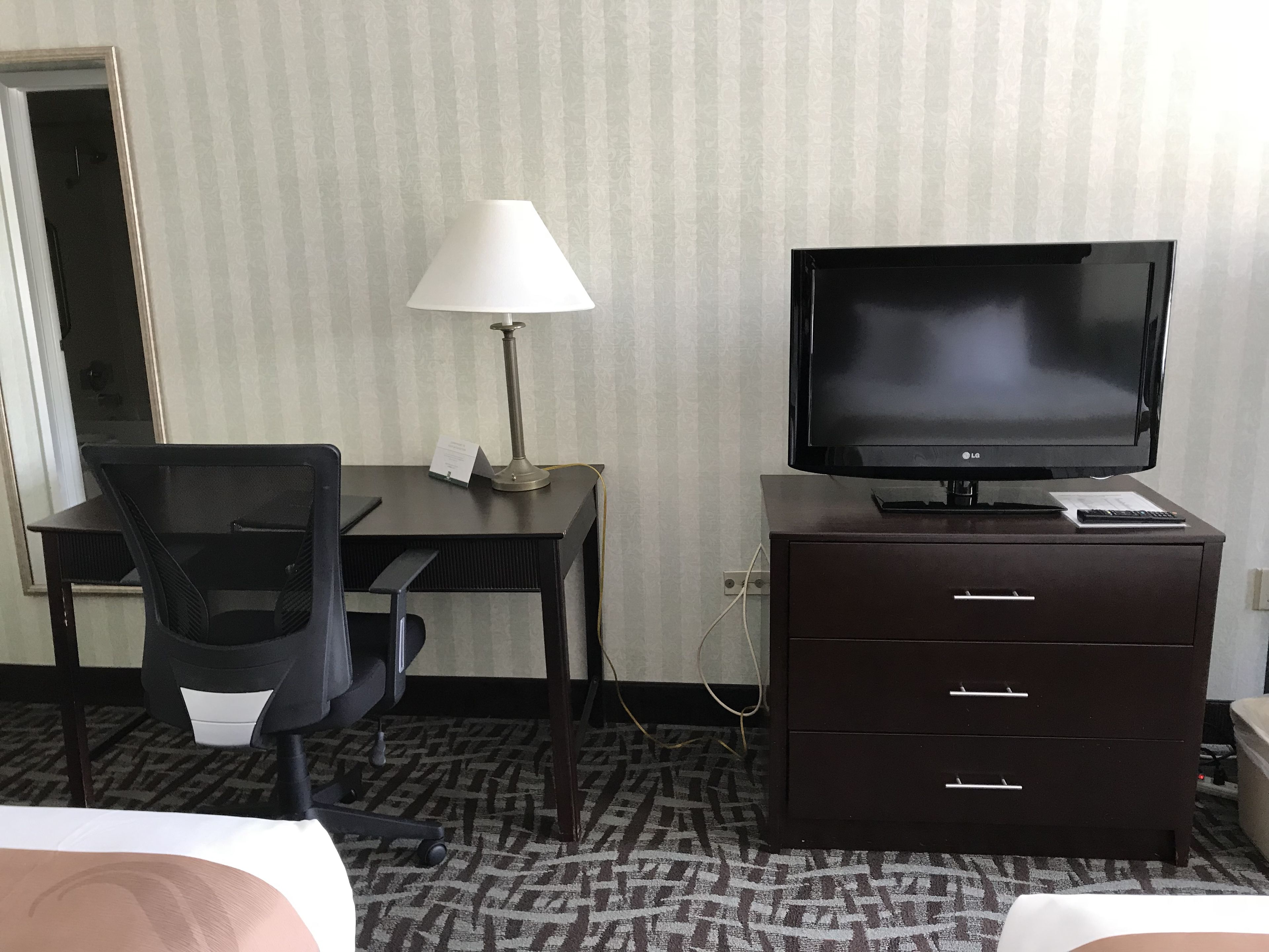 Ein Hotelzimmer mit Schreibtisch, Fernseher, Stuhl, Lampe und Kommode.