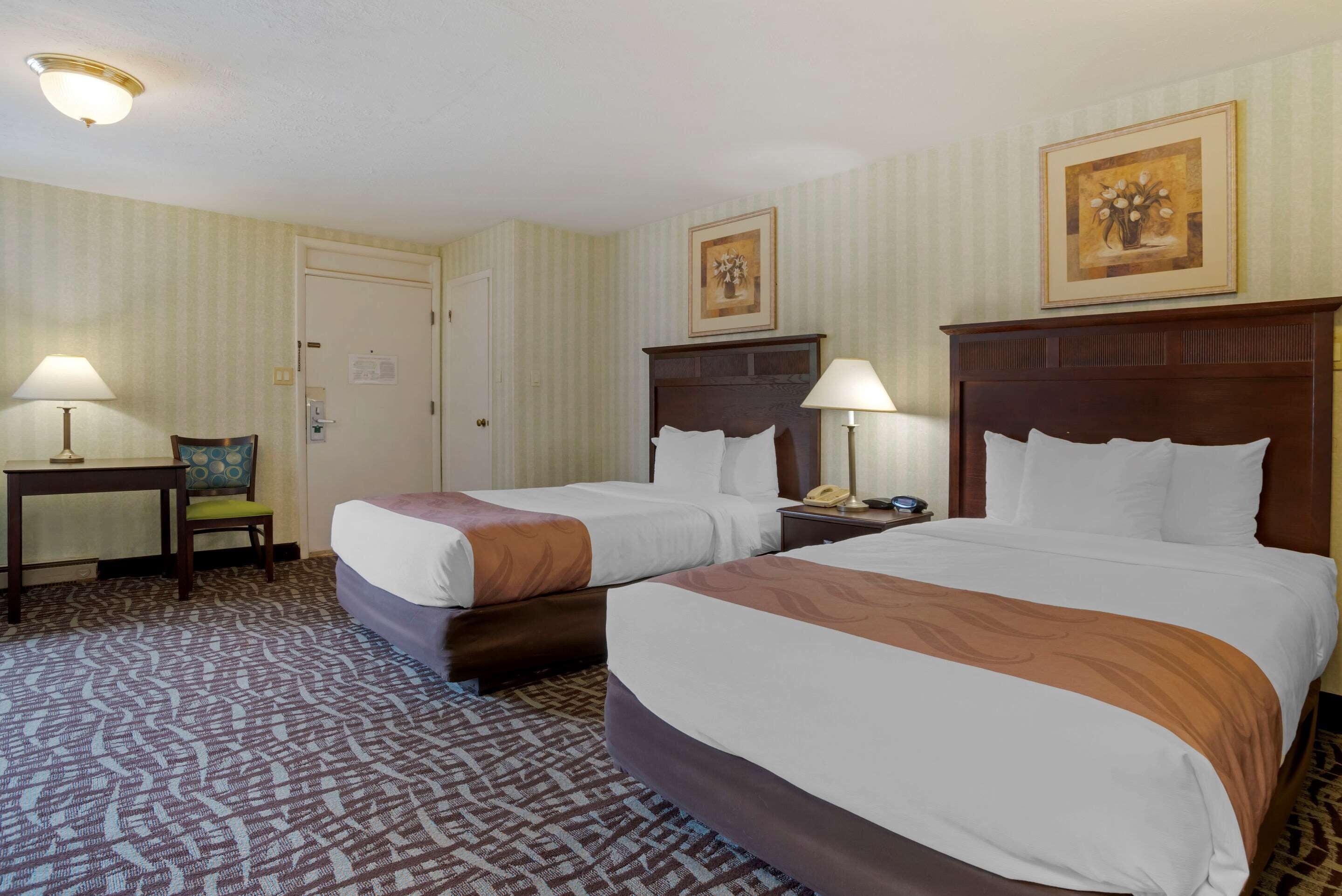 A hotel room with two beds, a desk, a chair, and a nightstand with a lamp.