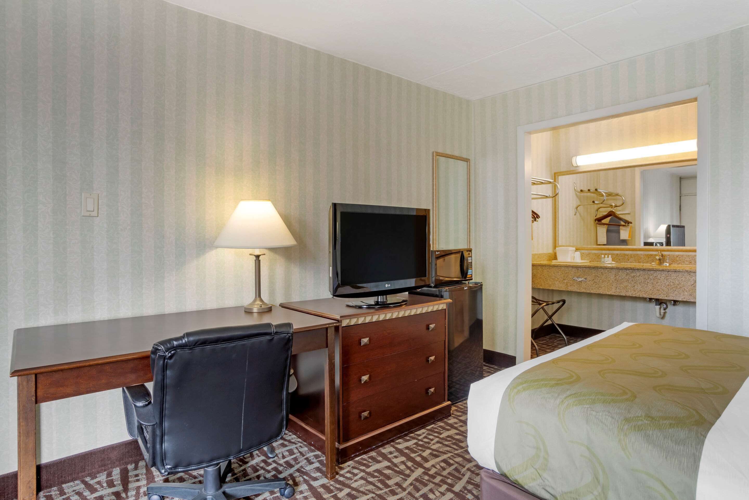 Habitación de hotel con cama, escritorio, televisor, silla, cómoda y espejo.