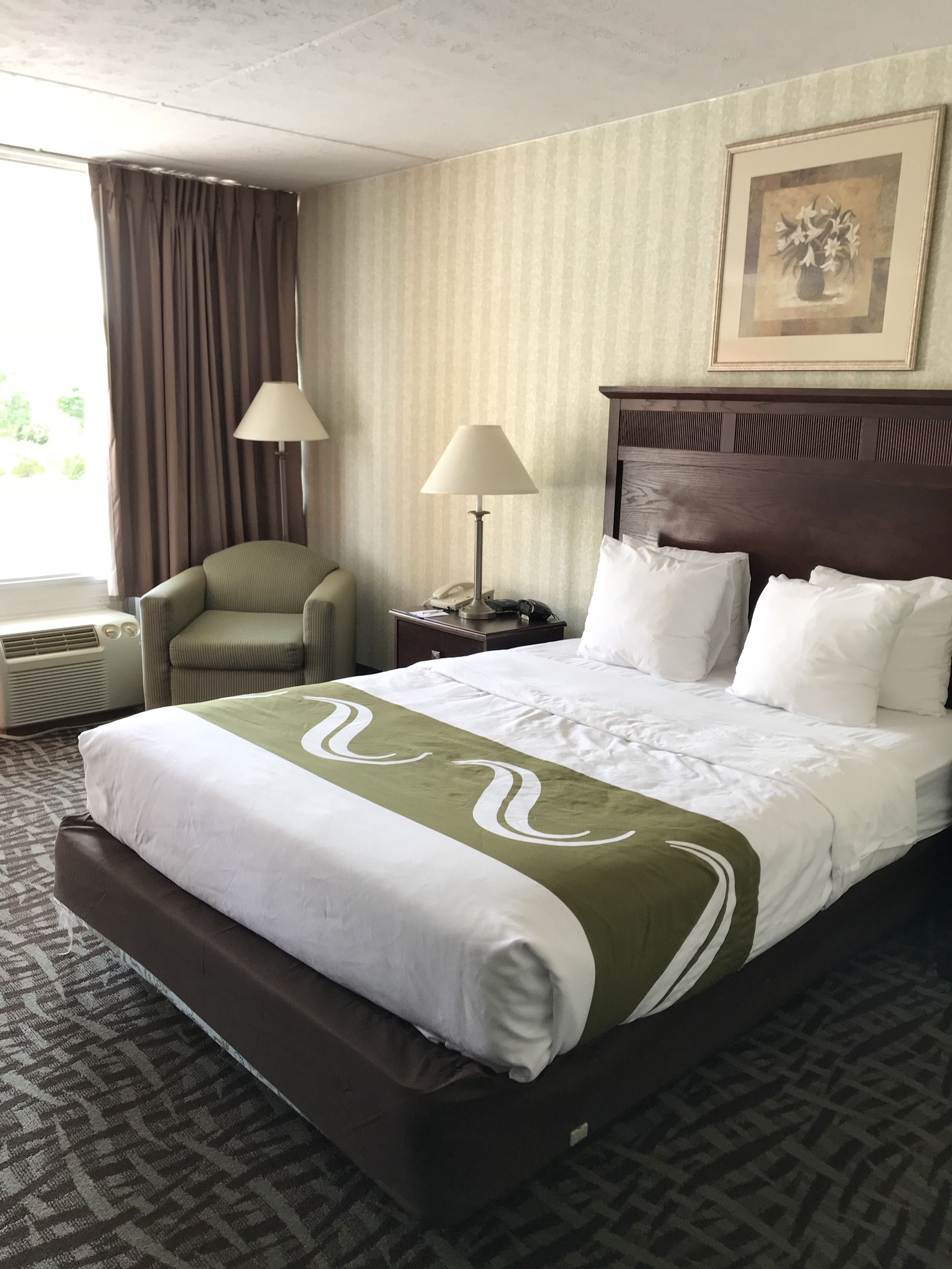 A hotel room with a large bed, a chair, a nightstand with a lamp, and a window with curtains.