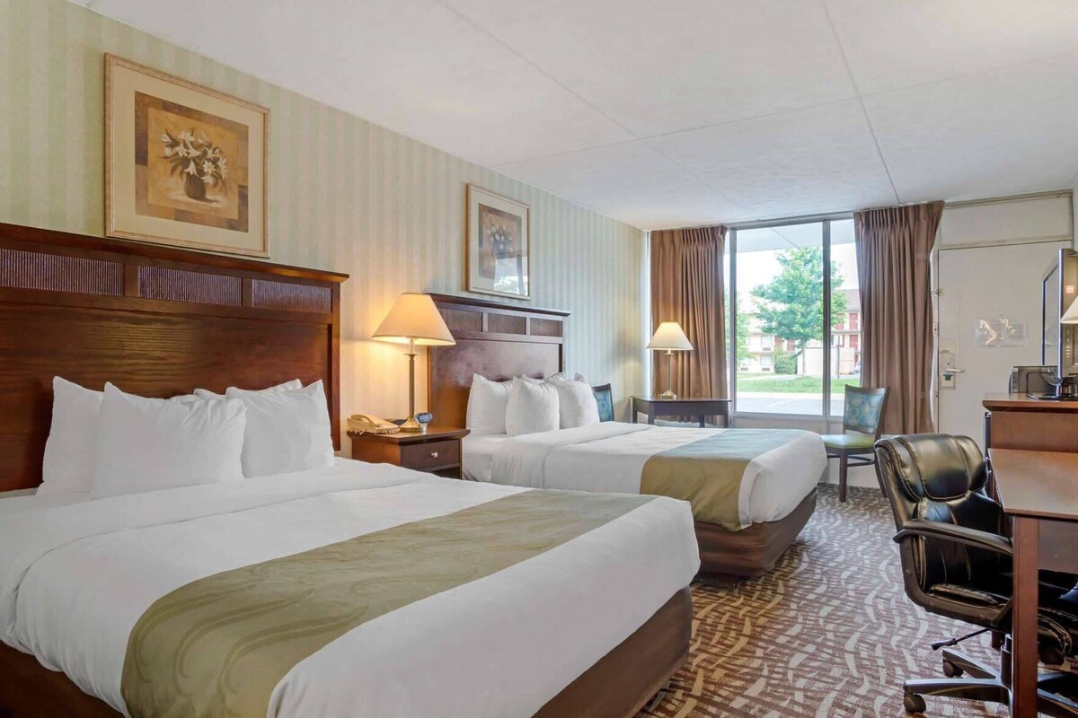 A hotel room with two beds, a desk, a chair, and a large window.