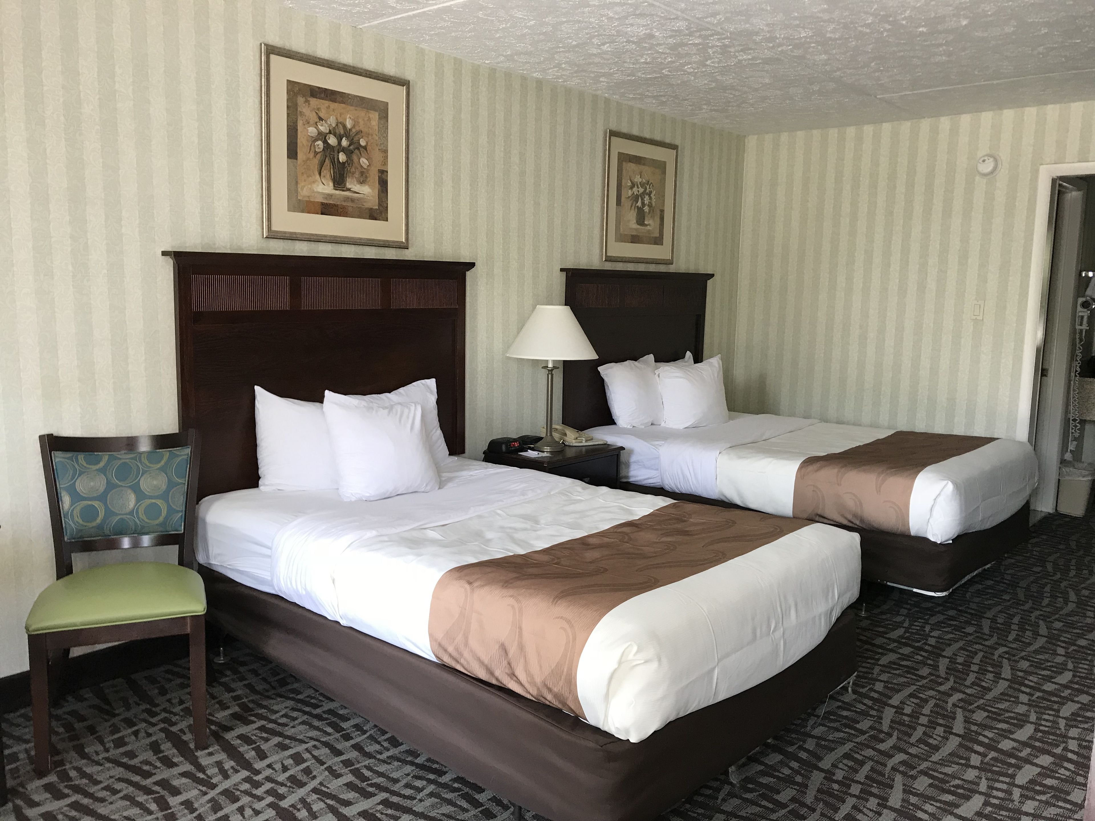 Habitación de hotel con dos camas, una silla, una mesita de noche y dos cuadros en la pared.