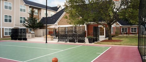 An outdoor basketball court with a green playing surface and a red border, surrounded by residential buildings and trees.