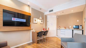 A hotel room with a flat-screen TV, a desk with two chairs, a kitchenette, and a seating area with a chair.