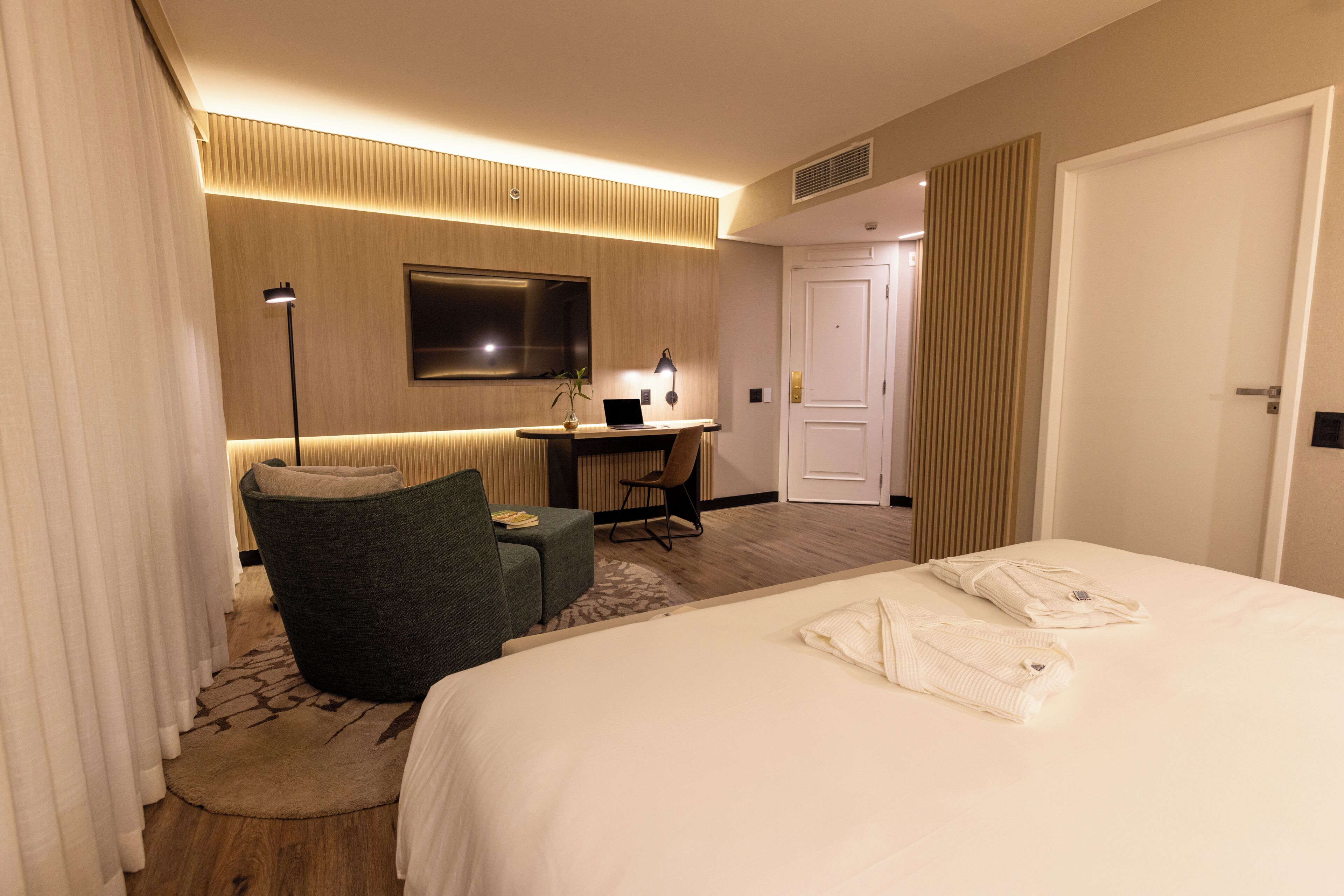 A hotel room with a bed, a desk with a laptop, a chair, and a large window.