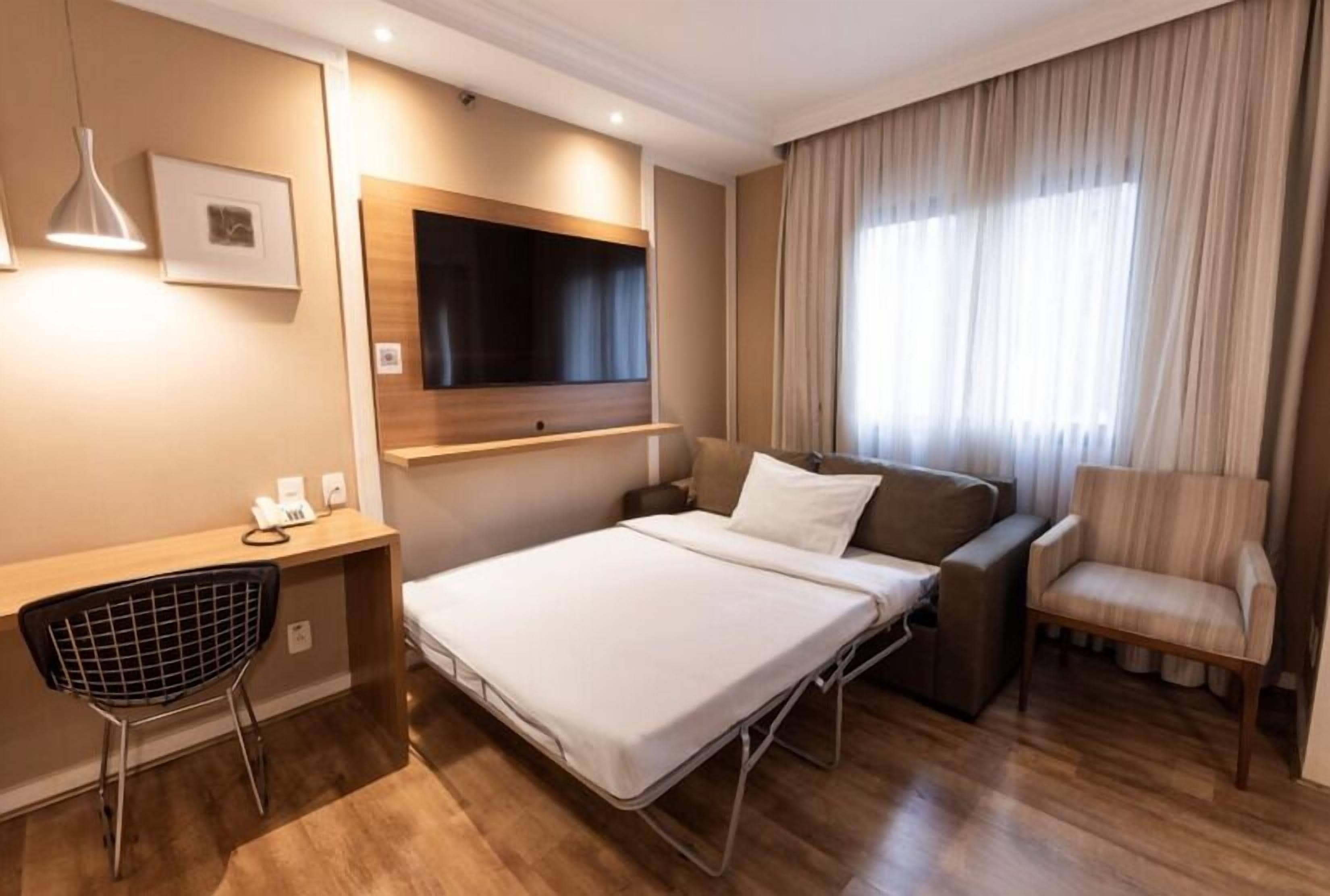 Ein Hotelzimmer mit Bett, Schreibtisch, Stuhl, Fernseher und einem Fenster mit Vorhängen.