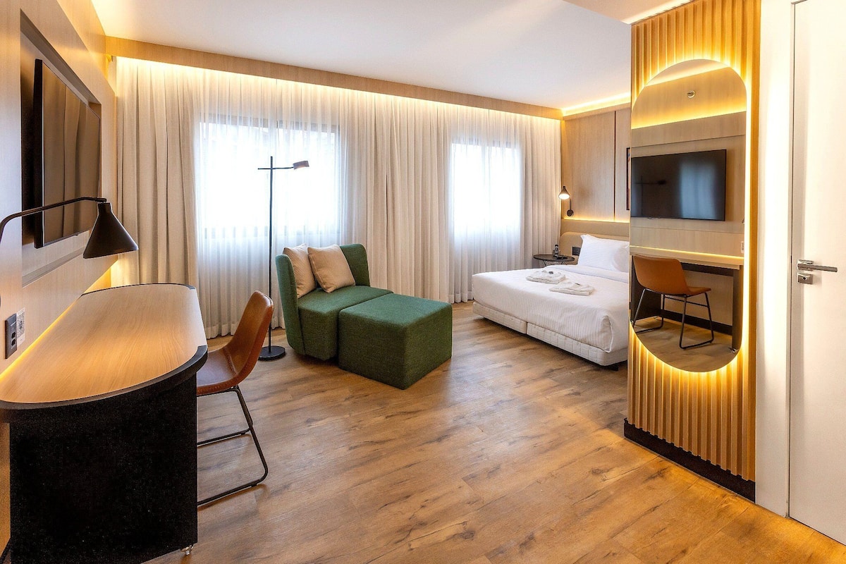Camera d'albergo con una scrivania, poggiapiedi verde, letto, televisore e tende.