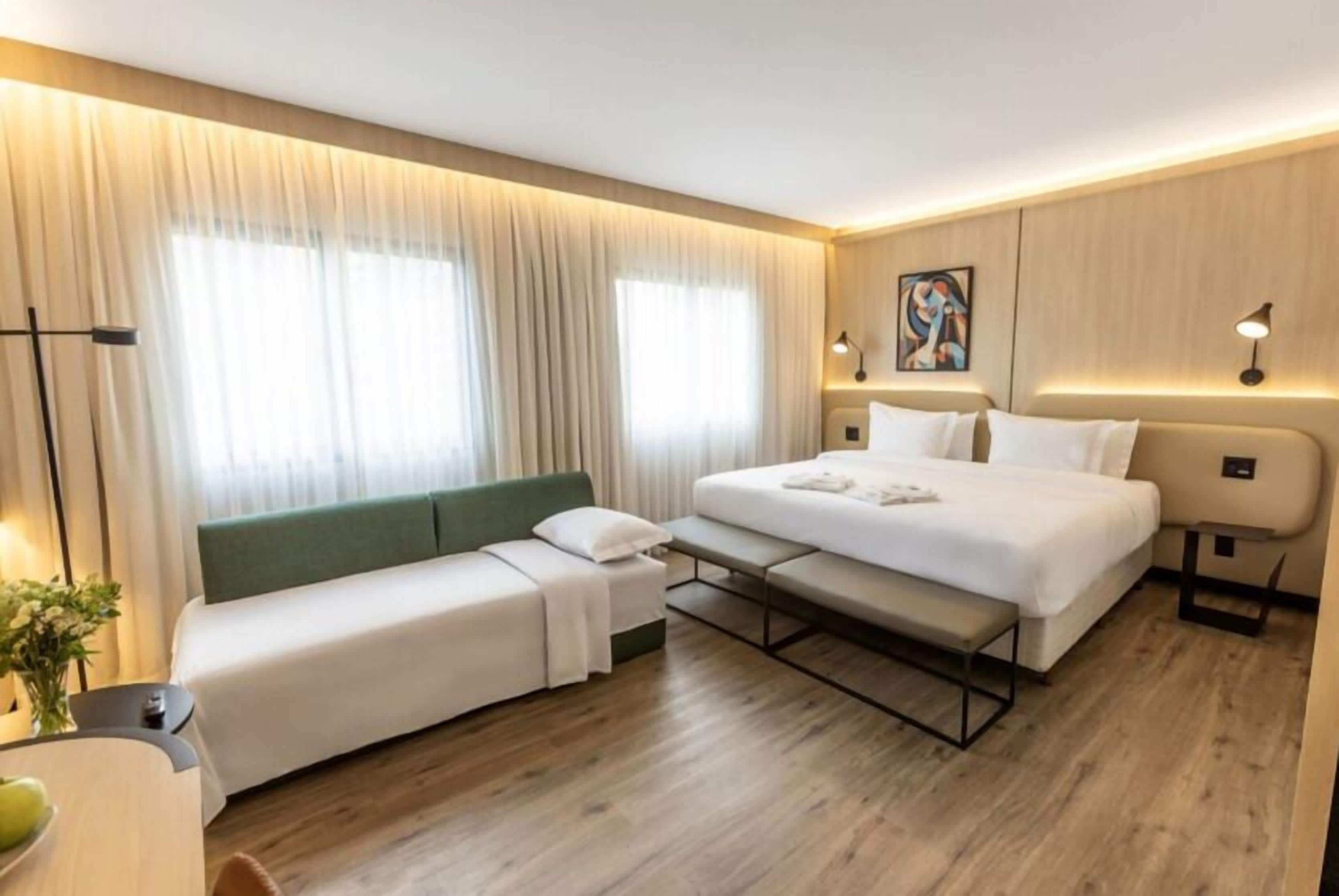 A modern hotel room with a bed, a sofa, a desk, and a large window with curtains.