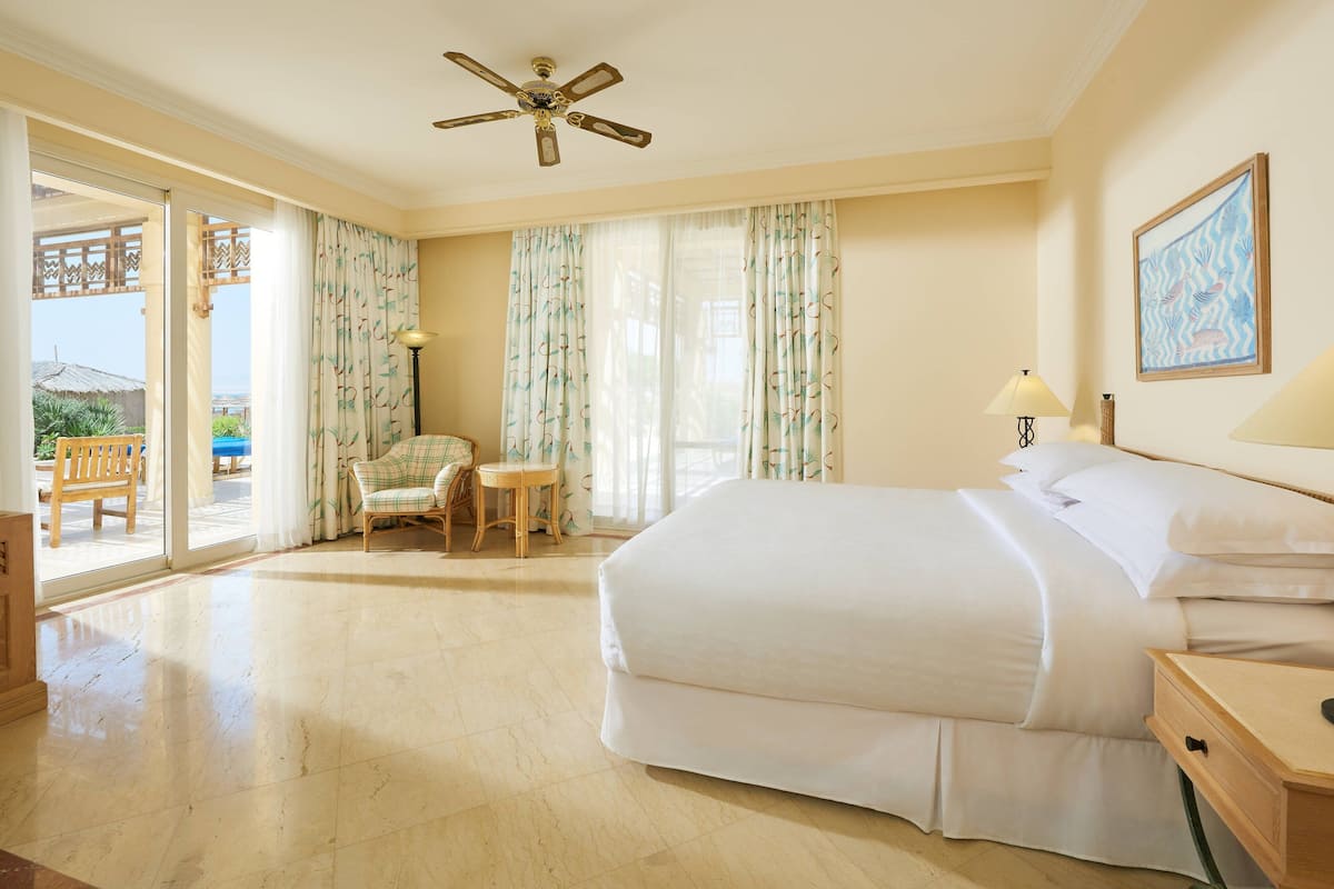 A hotel room with a large bed, a ceiling fan, a chair, a small table, and a view of the outside.