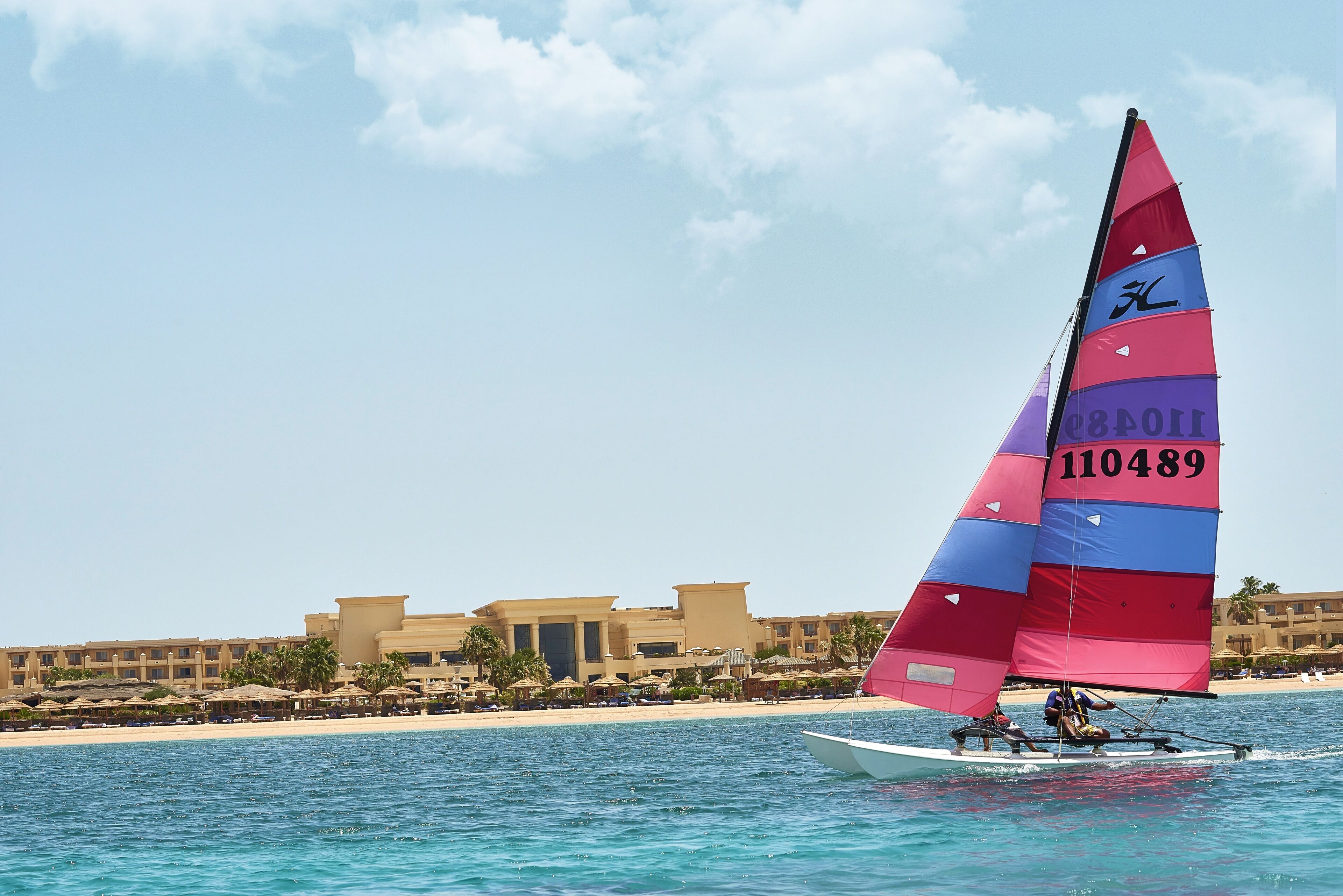 A sailboat with a colorful sail on a calm sea, with a resort and beach in the background.