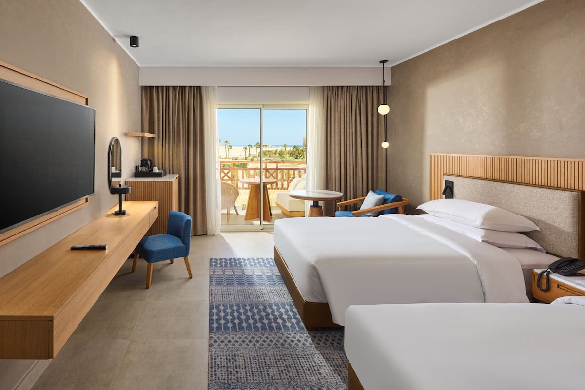 A hotel room with two beds, a desk with a chair, a TV, and a balcony with a table and chairs.