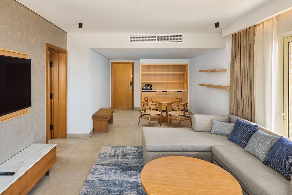 A modern living room with a sectional sofa, a round coffee table, a TV mounted on the wall, a dining area with a table and chairs, and built-in shelving.