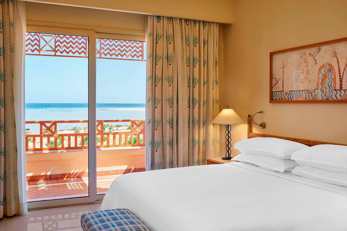 A hotel room with a bed, a nightstand, a lamp, and a view of the beach through a sliding door.