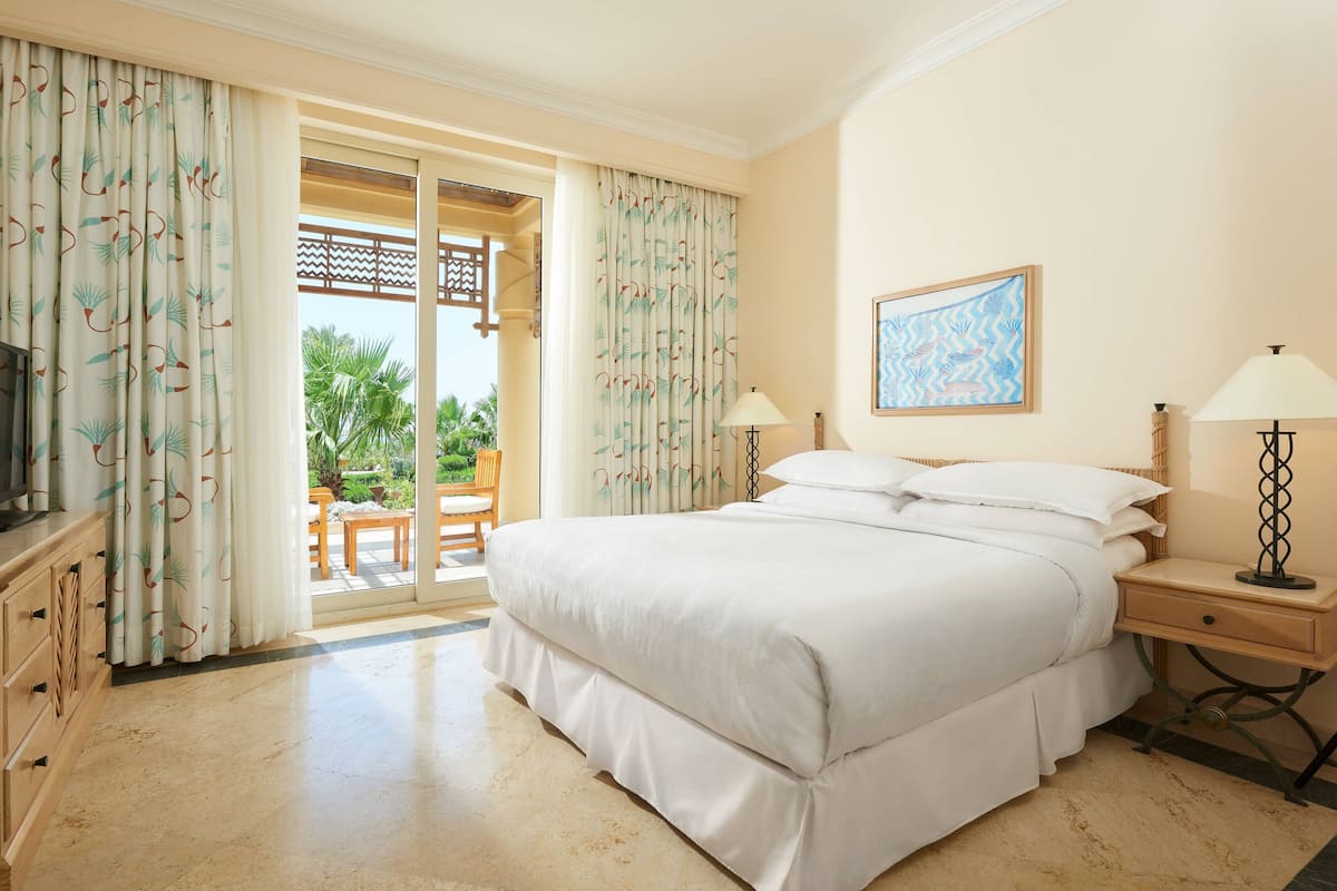 A hotel room with a large bed, two bedside tables with lamps, a television, and a sliding glass door leading to a balcony with outdoor furniture.
