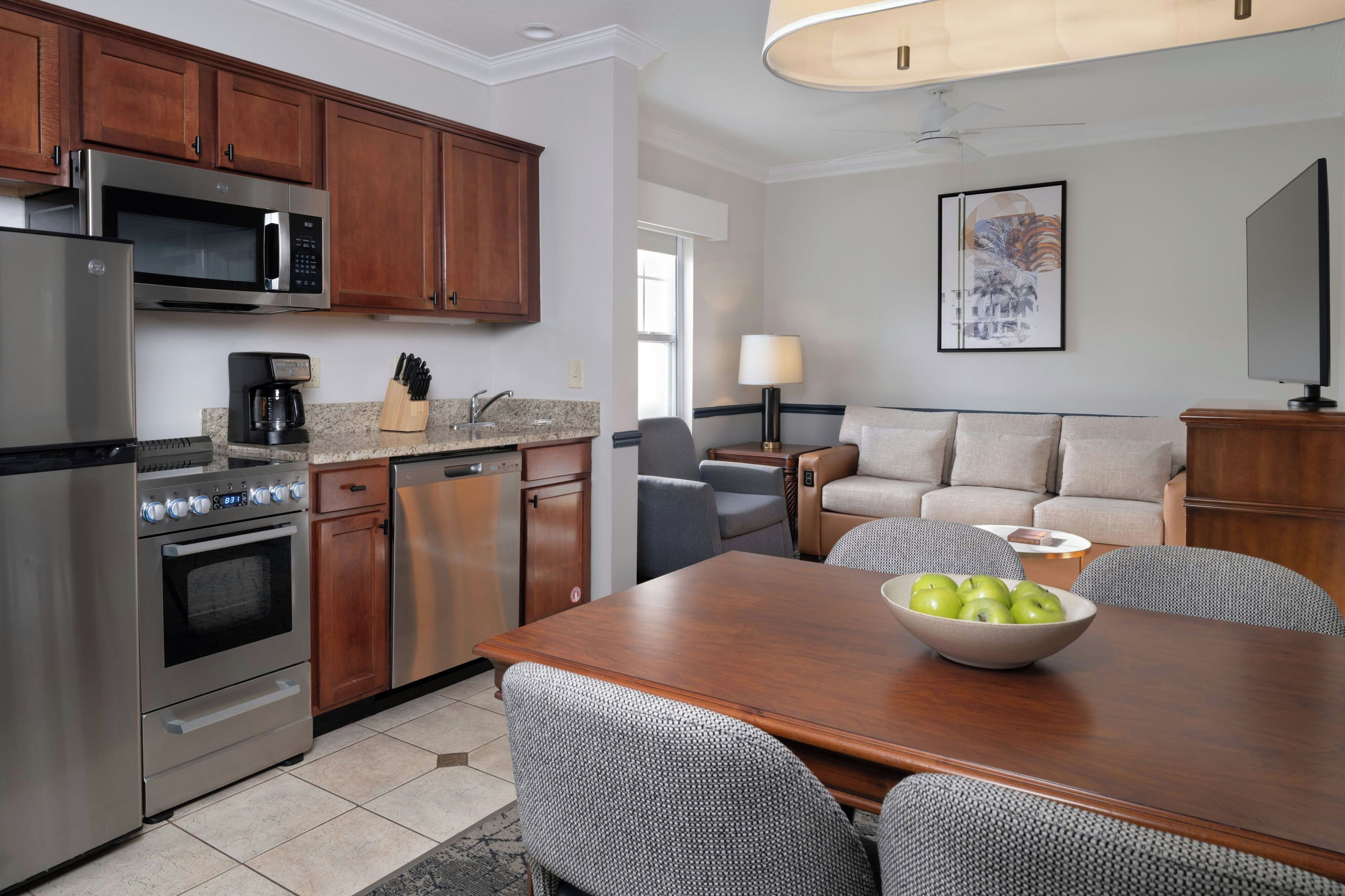 A modern kitchen with stainless steel appliances, a dining area with a wooden table and patterned chairs, and a living area with a sofa and a lamp.
