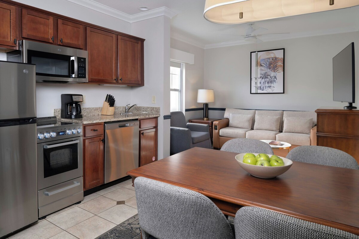 A modern kitchen with stainless steel appliances, a dining area with a wooden table and patterned chairs, and a living area with a sofa and a lamp.