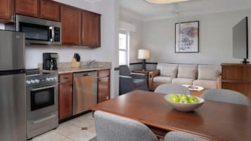 A modern kitchen with stainless steel appliances, a dining area with a wooden table and patterned chairs, and a living area with a sofa and a lamp.