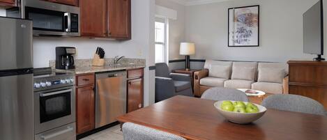 A modern kitchen with stainless steel appliances, a dining area with a wooden table and patterned chairs, and a living area with a sofa and a lamp.