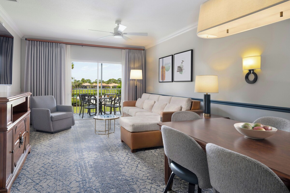 A hotel room with a sofa, armchairs, a dining table, and a view of the outdoors.