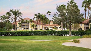 Campo de golf con césped cuidado, un búnker de arena y una hilera de edificios de varios pisos con balcones y ventanas amplias.
