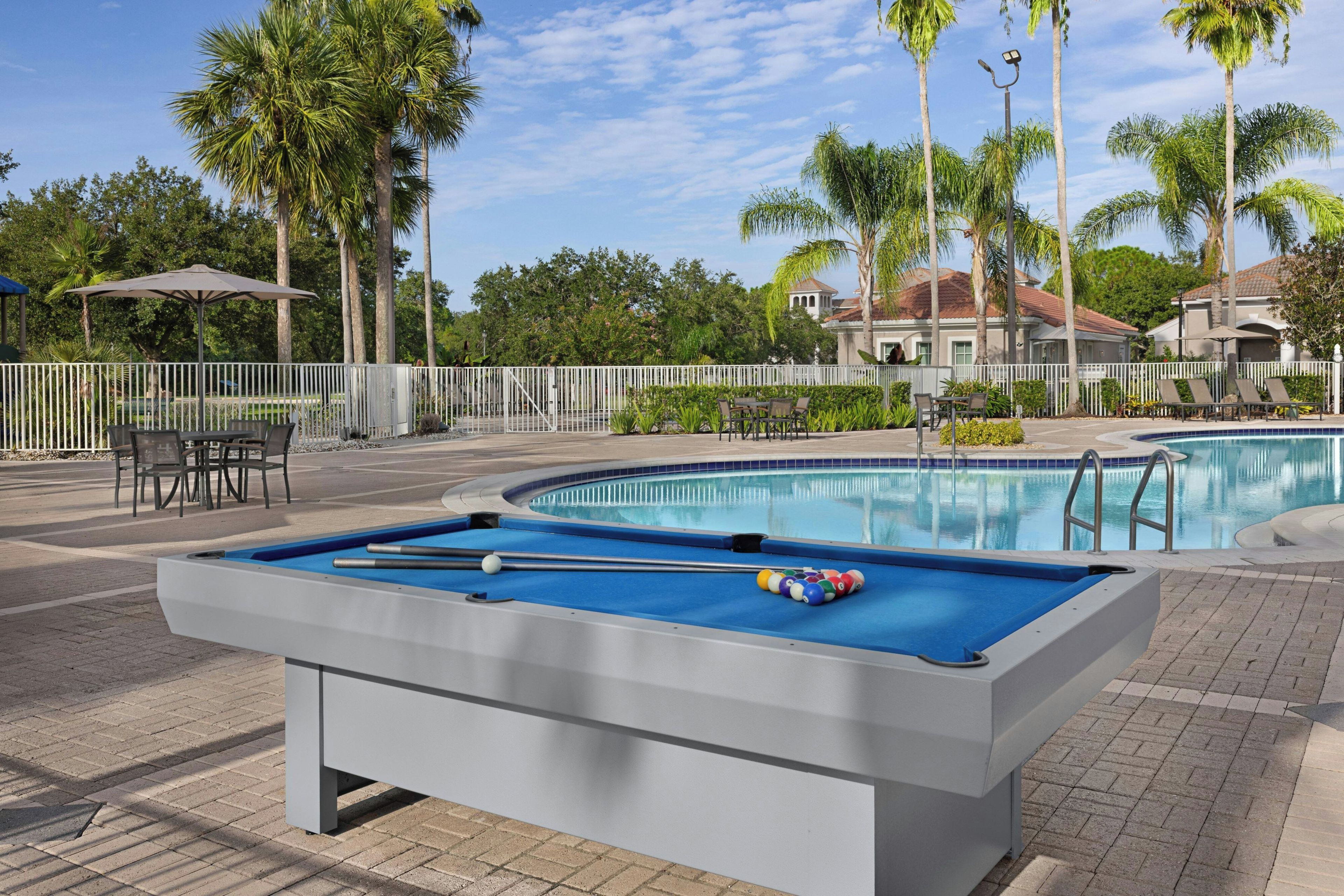 Mesa de bilhar com taco e bolas, área de estar com cadeiras e piscina.