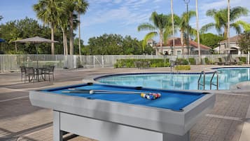 Mesa de bilhar com taco e bolas, área de estar com cadeiras e piscina.