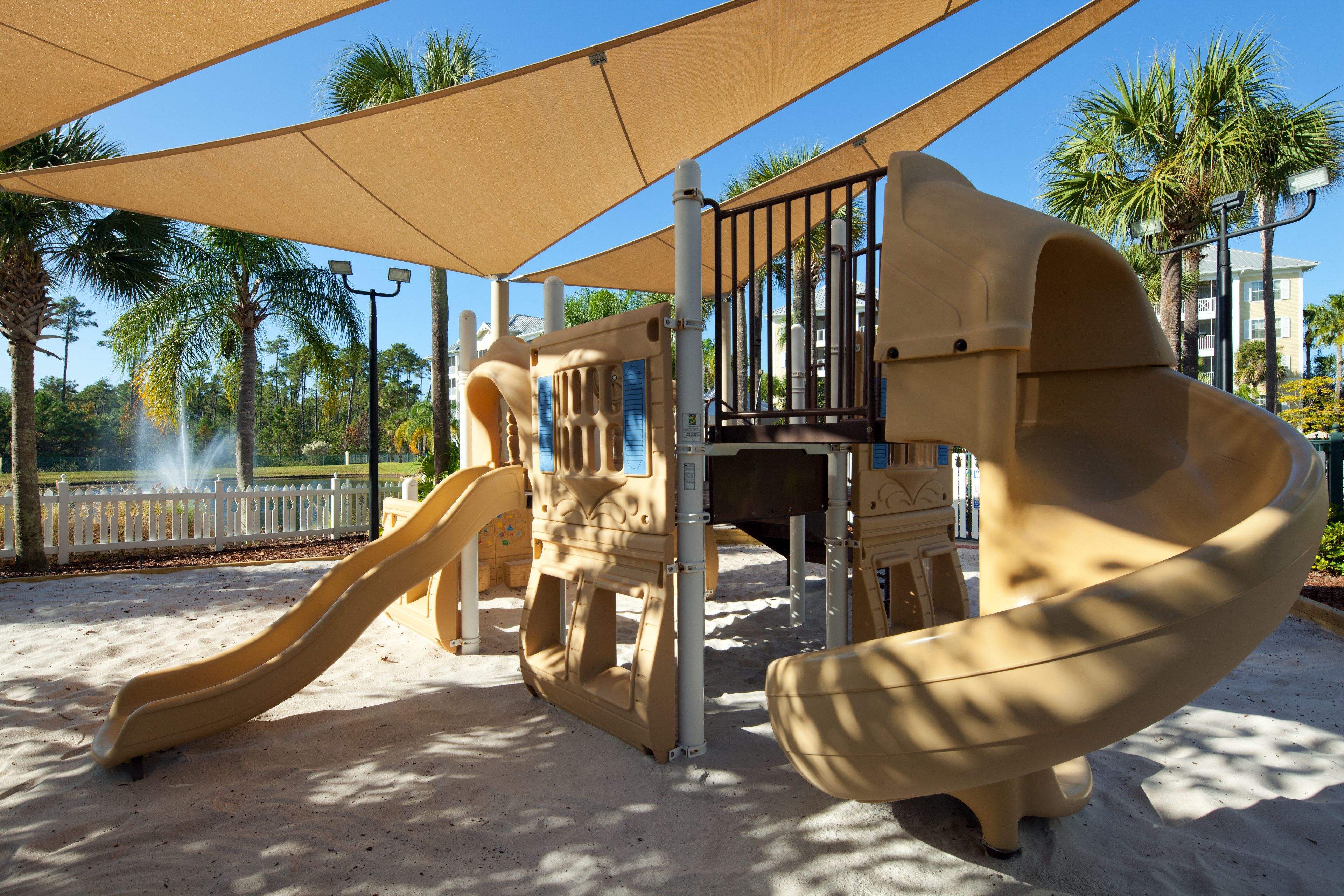 A playground with two slides and a climbing structure, surrounded by palm trees and a white fence.