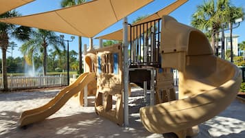 A playground with two slides and a climbing structure, surrounded by palm trees and a white fence.