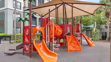 Un parque infantil con toboganes y estructuras para escalar, rodeado de una zona cubierta y palmeras.