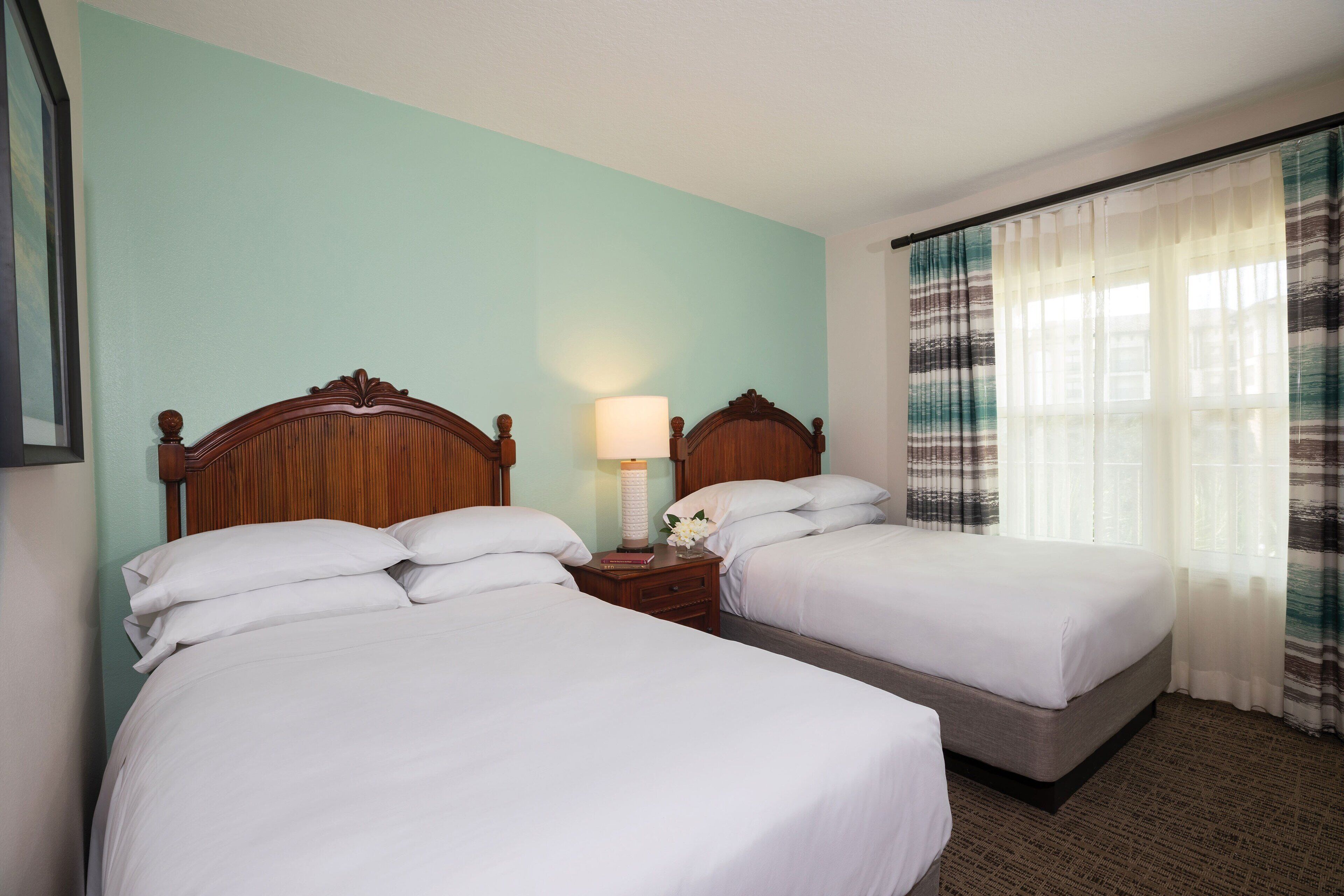 Une chambre d’hôtel avec deux lits simples, une table de chevet avec une lampe et une fenêtre avec des rideaux légers.