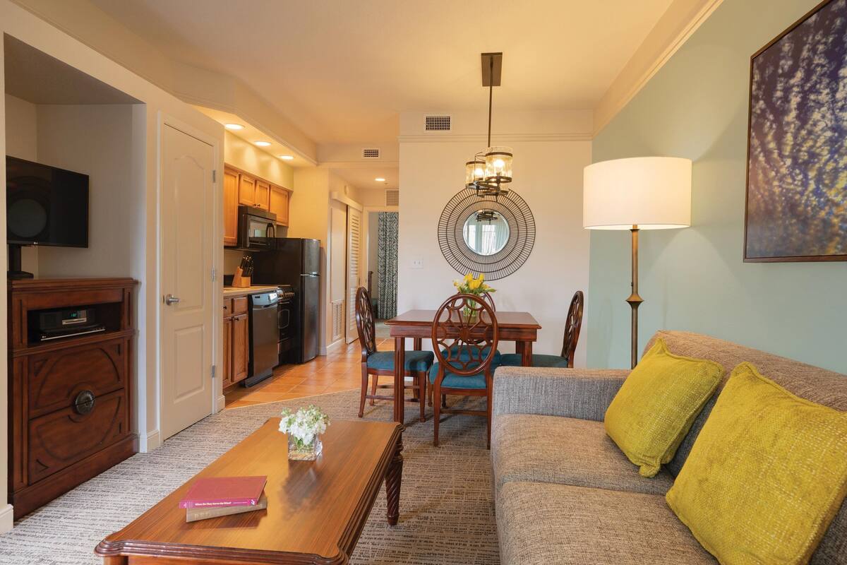 A living room with a sofa, coffee table, and a dining area with chairs and a table. There is a kitchen in the background with a microwave and refrigerator.