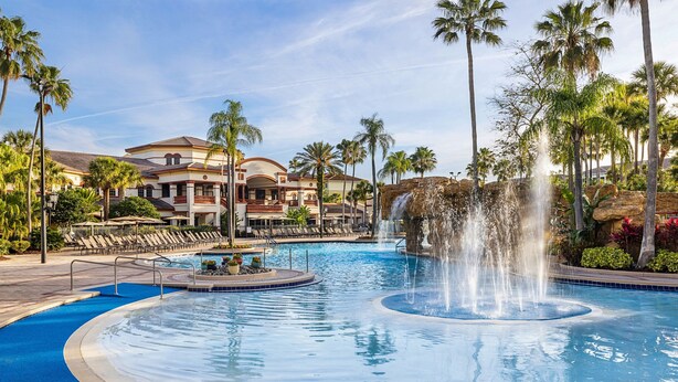 A resort pool with a central water feature, surrounded by palm trees and lounge chairs.