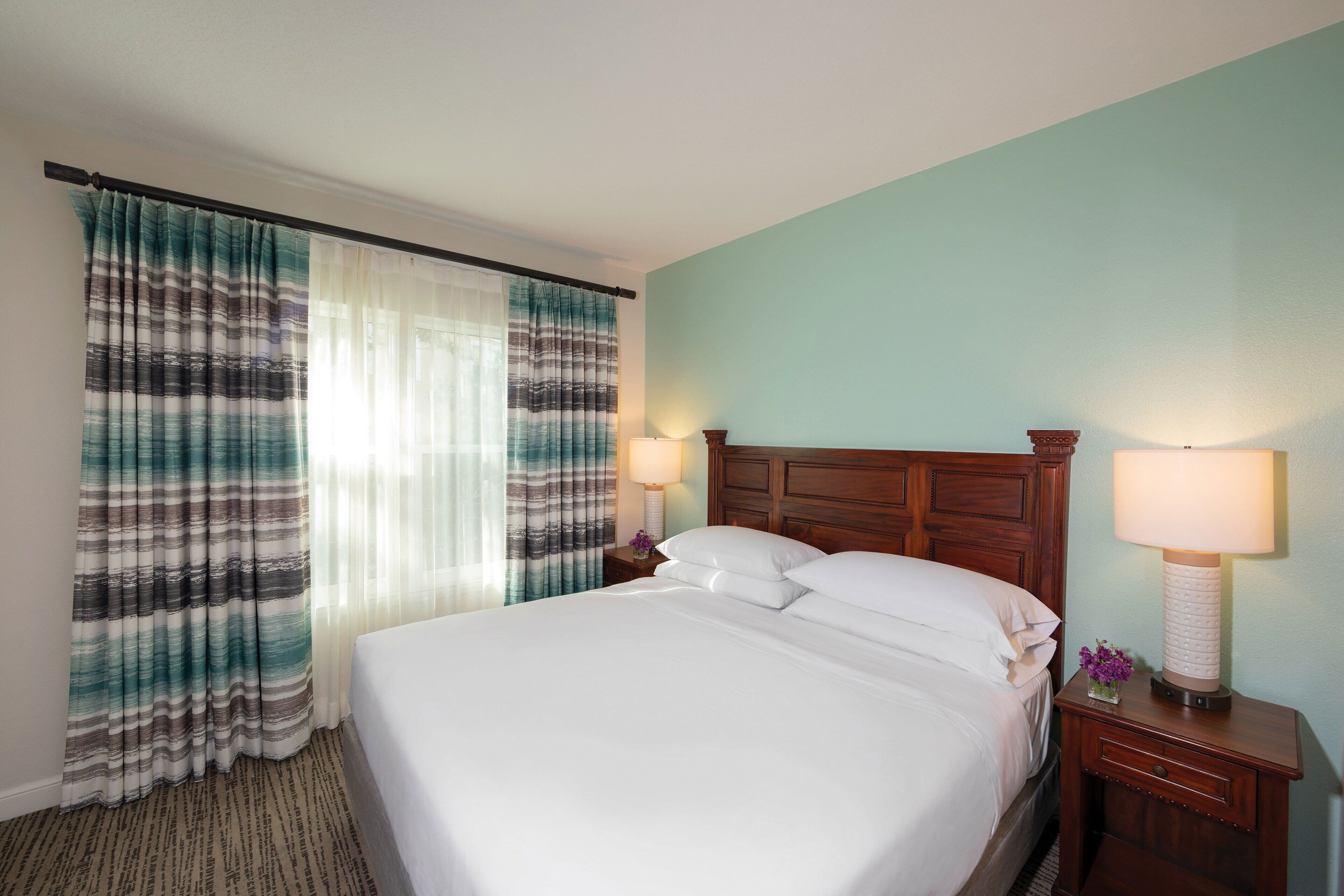 Una habitación de hotel con una cama grande, dos mesitas de noche con lámparas, un cabecero de madera y vistas al exterior a través de cortinas translúcidas.