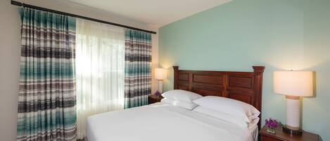 A hotel room with a large bed, two bedside tables with lamps, a wooden headboard, and a view of the outdoors through sheer curtains.