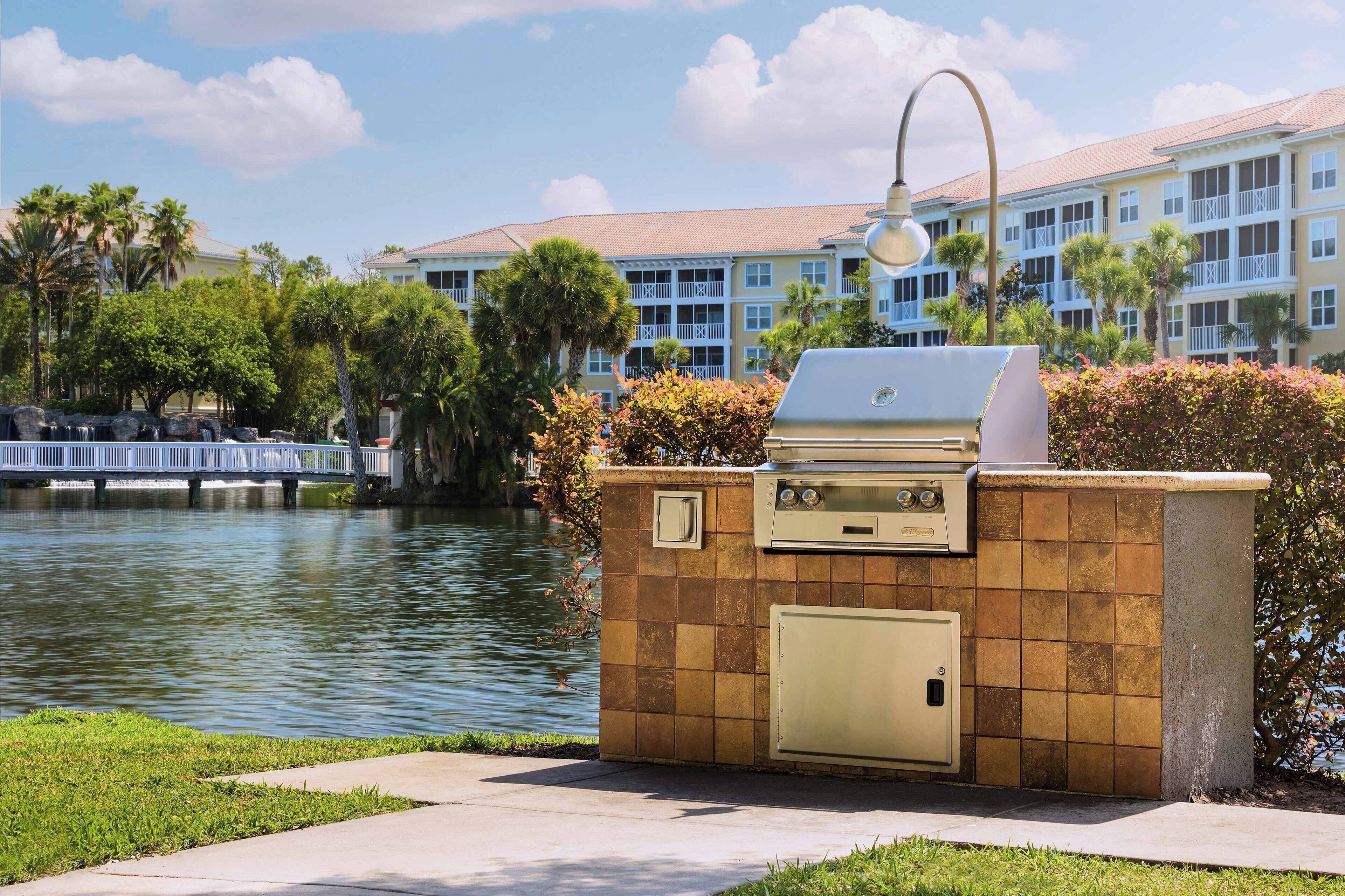Cocina exterior con parrilla y una lámpara, cerca de un cuerpo de agua y un edificio con balcones.