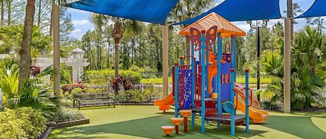 A playground with a slide and climbing structures, surrounded by palm trees and artificial turf.