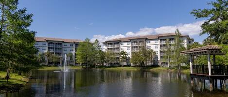 A tranquil lake with a fountain, surrounded by lush greenery and a multi-story building with balconies.