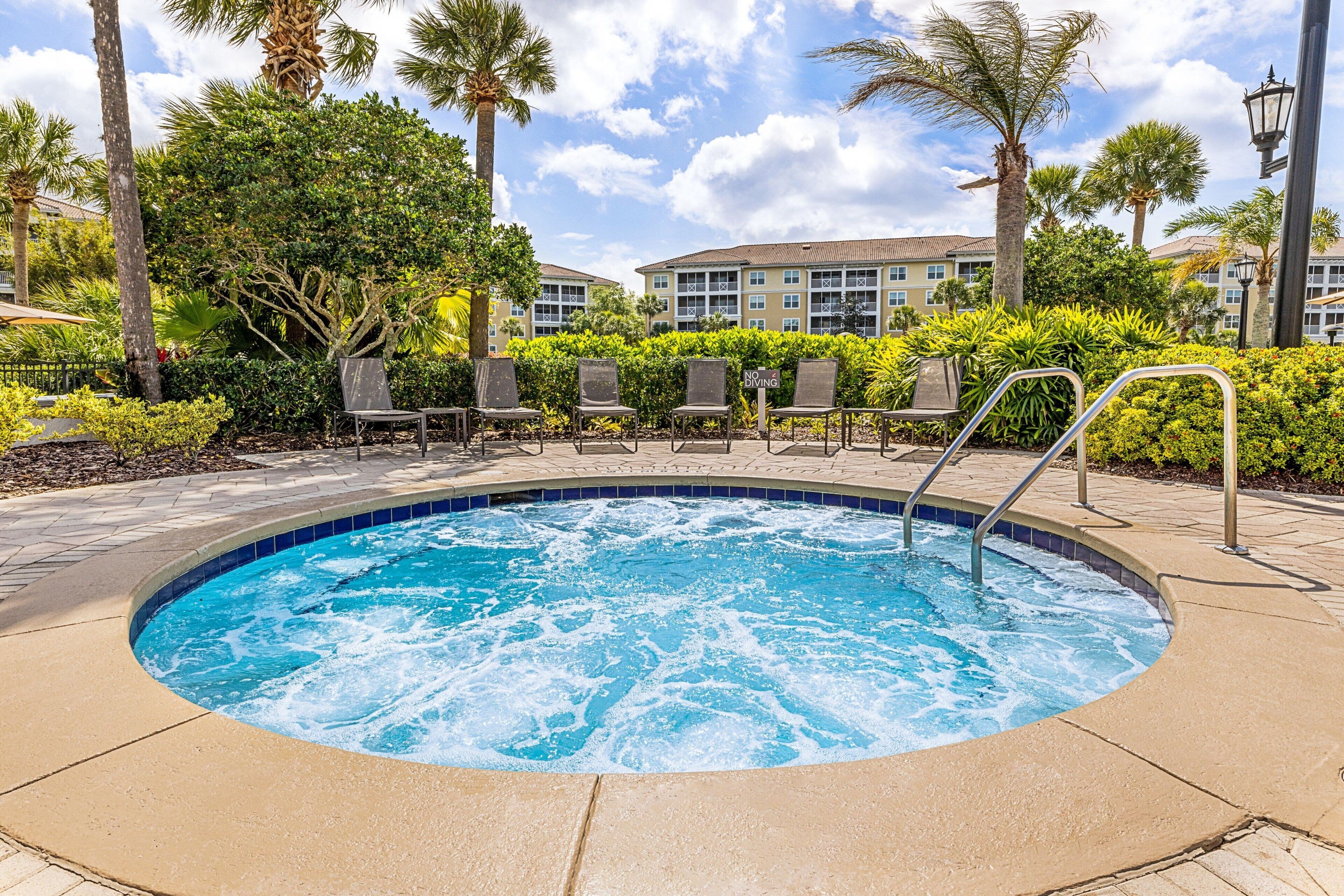 A hot tub surrounded by lounge chairs and palm trees, with a building in the background.