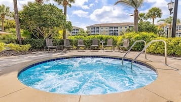 A hot tub surrounded by lounge chairs and palm trees, with a building in the background.