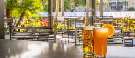 Un espace extérieur abrité avec des tables et des chaises, deux verres de bière et une vue sur des bâtiments et des arbres.