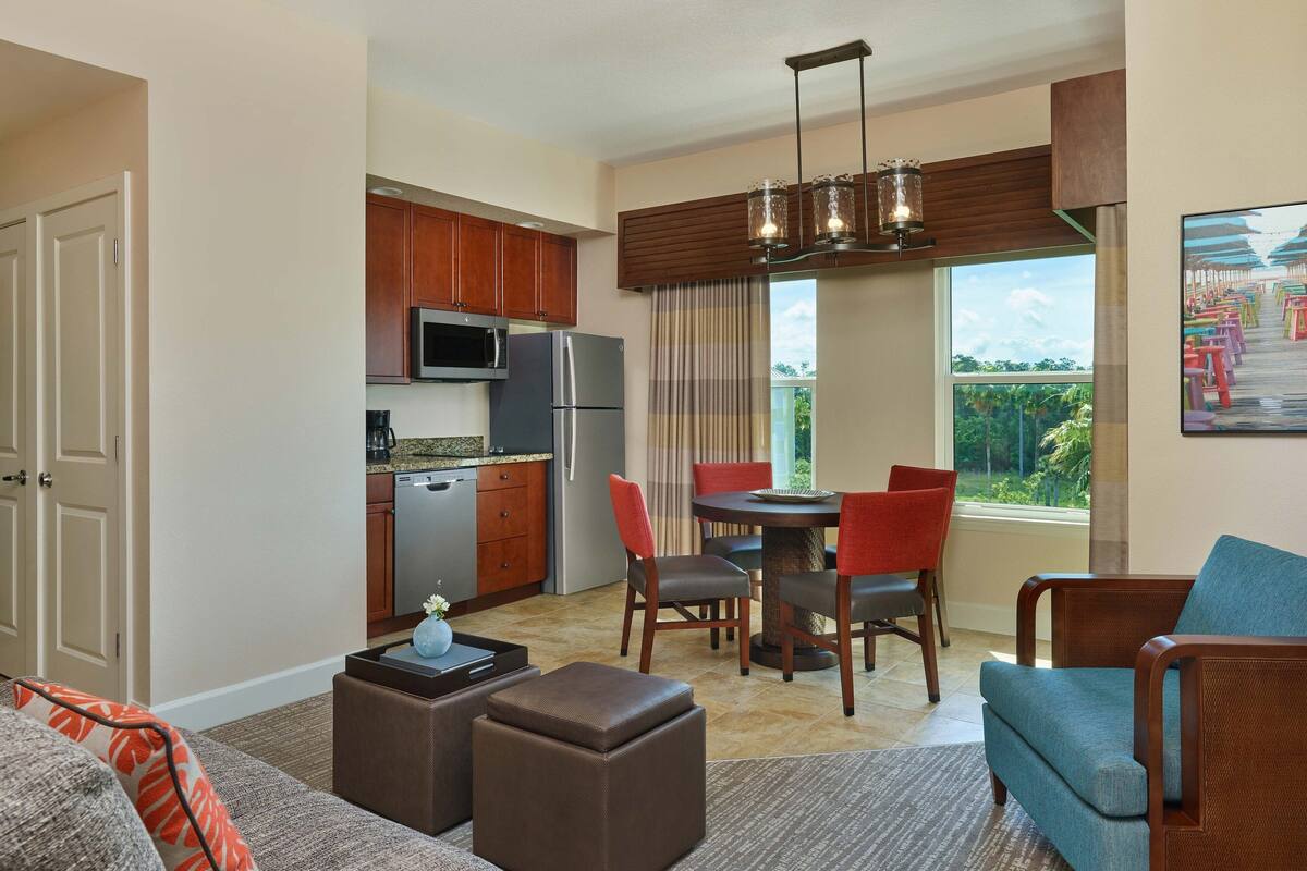 A modern kitchen with built-in appliances, a dining area with a round table and chairs, and a living room with a sofa and ottoman.