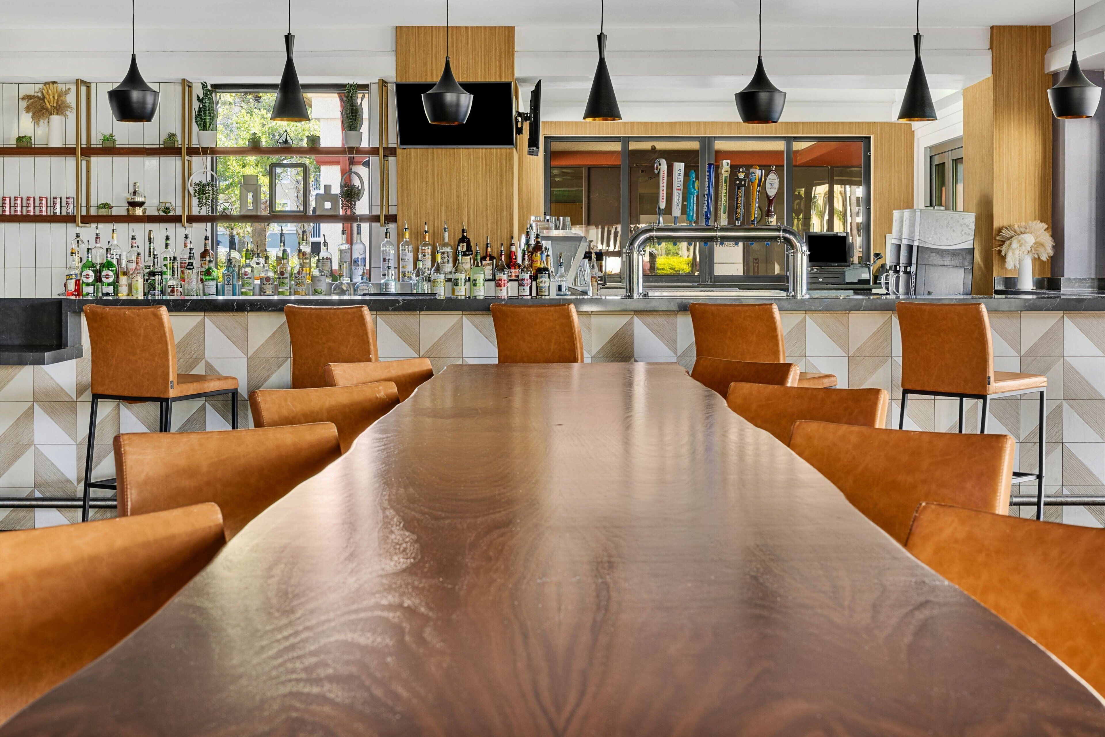 Une longue table en bois, entourée de chaises en cuir marron, dans un espace bar moderne agrémenté d’un mur recouvert de carreaux et de luminaires suspendus.