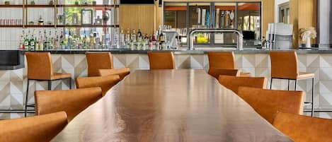 Une longue table en bois, entourée de chaises en cuir marron, dans un espace bar moderne agrémenté d’un mur recouvert de carreaux et de luminaires suspendus.
