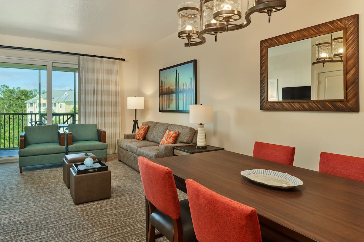 A hotel room with a dining table, red chairs, a sofa set, a lamp, a painting, and a large mirror.